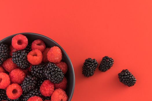 Blackberries and raspberries in a black bowl. Red background. Top view.