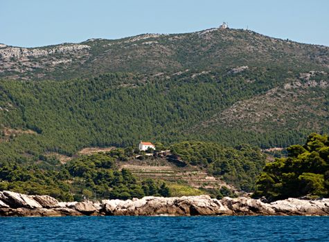 This is a picture of a chuch on a hill; surronded by sea; trees and mountains and visible sky-strip