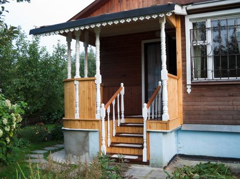 The pictures shows a new wooden porch in a country house, surrounded by green trees and bushes