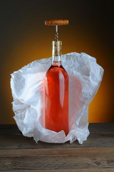 A bottle of blush wine wrapped in tissue paper, on a rustic wood surface and a light to dark warm background.
A vintage cork screw is inserted in the bottle.