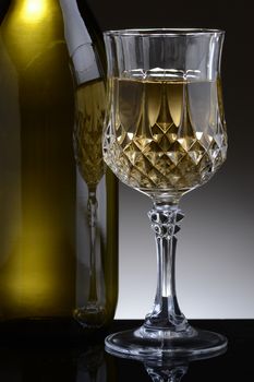 Closeup of a cut crystal glass of white wine and bottle against a light to dark gray background.