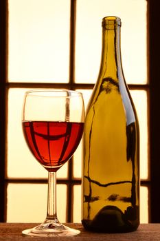 A wine still life with warm tones. One empty wine bottles and a glass of red wine in front of a window with warm sunlight.
