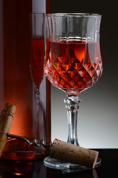 Closeup of a cut crystal glass of red wine and bottle against a light to dark gray background.