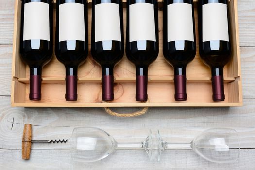 Overhead shot of a case of red wine bottles with blank labels  on a rustic white wood table with wine glasses and cork screw below. Horizontal format.