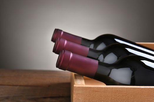 Three bottles of red wine in a wood crate with a light to dark gray background.