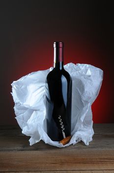 Red Wine Bottle and Cork Screw with tissue paper wrapping on wood surface and light to dark background.