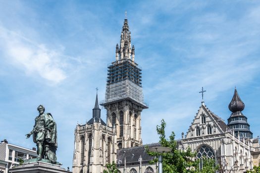 Antwerpen, Belgium - June 23, 2019: Bronze Peter Paul Rubens statue fronts towers and nave of Notre Dame, Onze-Lieve-Vrouw, cathedral against light blue sky. Some green foliage.
