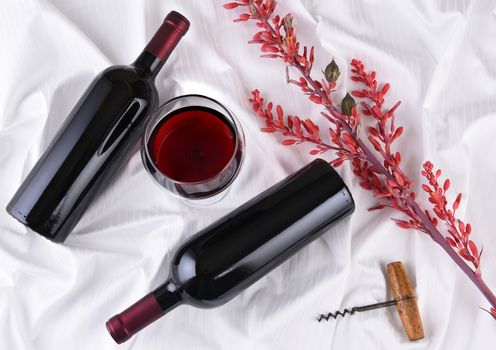 Wine glass in the center of two bottles laying on their sides on white fabric surface with corkscrew and flowers. Horizontal orientation.