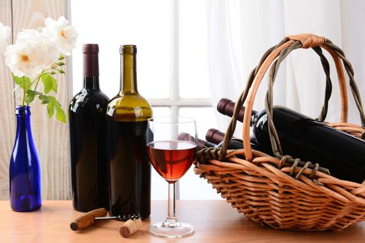 Wine still life with a basket of bottles and a glass of red wine and white roses in a vase on a table in front of a window with curtains. Horizontal format.