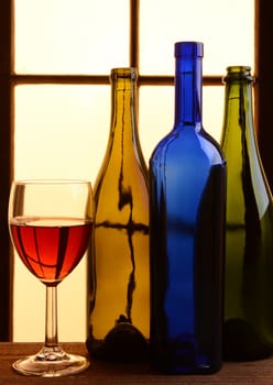 A wine still life with warm tones. Three different wine bottles and a glass of red wine in front of a window with warm sunlight.