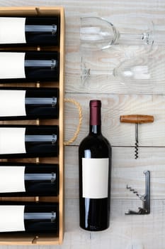 High angle shot of a case of red wine bottles next to a single bottle two wineglasses and cork screws on a rustic white wood table. Vertical format.