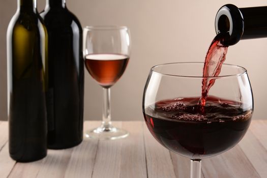 Closeup of a glass of wine and a bottle pouring. Out of focus wine bottles and another wineglass are in the background. Horizontal still life with warm tones and light to dark background lighting.