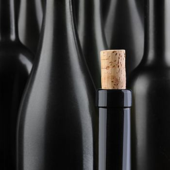 Closeup of a wine bottle with the cork partially out, in front of an out of focus group of larger bottles. Selective focus and color. Square Format.