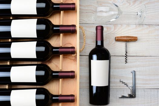 High angle shot of a case of red wine bottles with blank labels next to a single bottle a wineglass and cork screws on a rustic white wood table. Horizontal format.