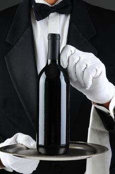 Closeup of a waiter wearing a tuxedo and white gloves holding a wine bottle on a serving tray. Vertical format. The man is unrecognizable.