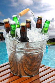 Close up of a beer bucket on poolside teak table. Swimming pool with beach balls and house in background. Cloudy blue sky and ocean in far background.