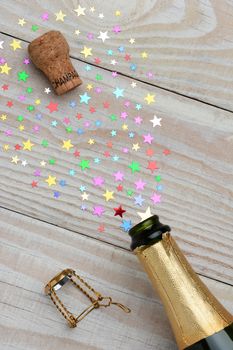 Overhead of a bottle of champagne with the cork popping on a rustic wood table. The spray from the bottle is in the form of colorful stars of various sizes. With copy space it is perfect for New Years projects.