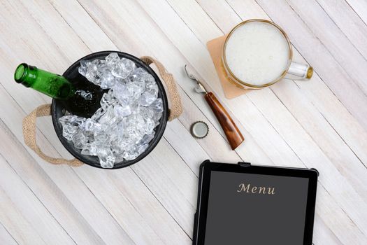 Overhead shot of an ice bucket with an opened beer bottle, a mug of beer and tablet computer for electronic ordering on a rustic white wood table. Horizontal format with copy space.