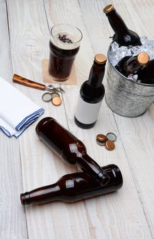 High angle shot of a party bucket filled with beer bottles on a rustic wood table. Empty bottles and bottle caps are on the table along with a glass of dark ale and bar towel.