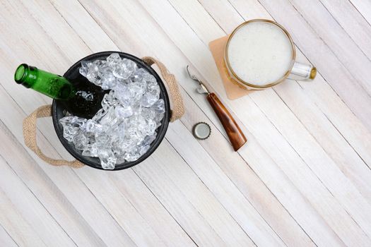 Overhead shot of an ice bucket with an opened beer bottle, a mug of beer and opener on a rustic white wood table. Horizontal format with copy space.