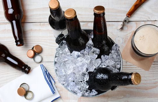 High angle shot of a glass of beer with a metal bucket with unopened bottles. The rustic wood table has empty bottles and caps and an bottle opener. Horizontal Format.