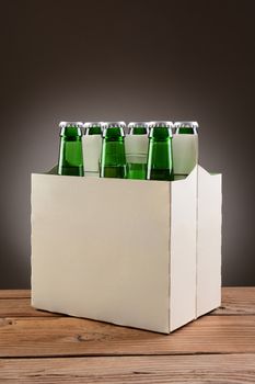 Closeup of a six pack of green beer bottles on a rustic wooden table. Vertical format with a light to dark gray spot background.