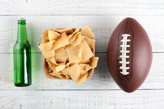 Beer bottle, bowl of chips and an American style football on a rustic whitewashed wood surface. Horizontal format. The bottle is without label.