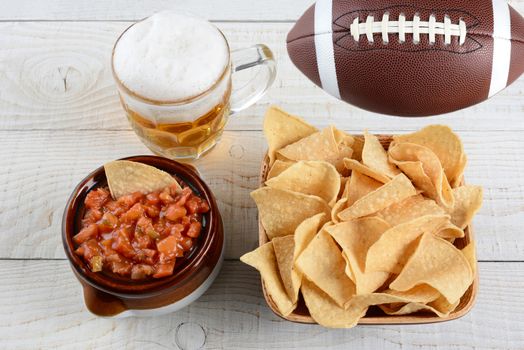 High angle shot of a bowl of corn chips a crock full of fresh salsa a mug of beer and an American football on a whitewashed rustic wood table. Horizontal format. 