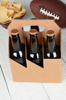 High angle shot of a six pack of beer bottles a bowl of chips and an American football. Vertical format with focus on the beer bottles. Bottles have no labels and the carrier is blank.