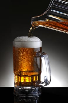 Closeup of a beer mug being filled from a pitcher pouring cold ale into the glass. Vertical format on a light to dark gray background.
