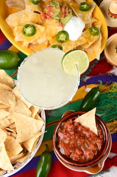 Overhead view of a margarita cocktail surrounded by nachos, chips, salsa on a bright Mexican, table cloth. Vertical format. Cinco de Mayo theme.