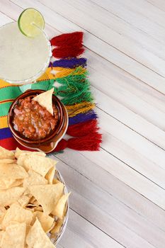 Overhead view of a margarita cocktail with chips and salsa on a white rustic wood table. Vertical format with copy space.