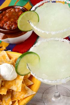 High angle view of two margarita cocktails for a Cinco de Mayo celebration. Surrounded by nacho chips and salsa on a bright Mexican table cloth.