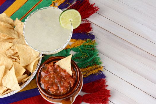 High angle view of a margarita cocktail with chips and salsa on a white rustic wood table. Horizontal format with copy space.