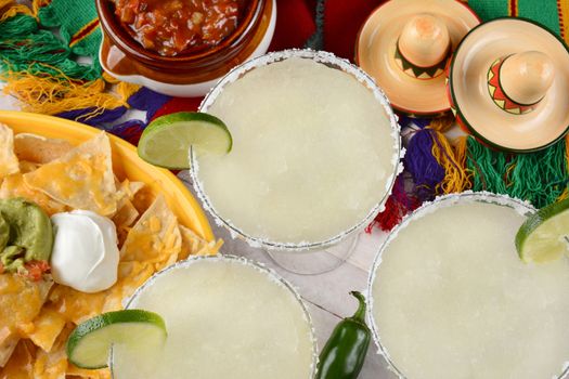 High angle view of three margarita cocktails surrounded by nachos, chips and salsa on a bright Mexican, table cloth. Horizontal format. Perfect for Cinco de Mayo projects.