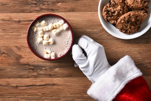 High angle closeup of Santa Claus holding a mug of hot cocoa on a wood table with plate of cookies.
