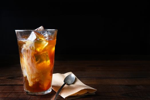 Closeup of a glass of iced coffee on a dark wood table. Fresh poured cream is permeating through the glass with a napkin and spoon lay next to the glass. Horizontal with copy space.