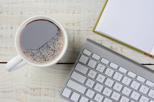 Overhead shot of a fresh brewed cup of coffee, an open book and a computer keyboard on a rustic white wood desk. Horizontal format.