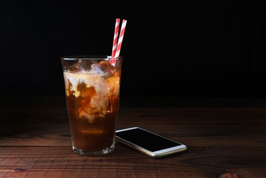 A glass of iced coffee with straws on a dark wood table. Fresh poured cream is permeating through the glass with a cell generic phone laying next to the glass. Horizontal with copy space.