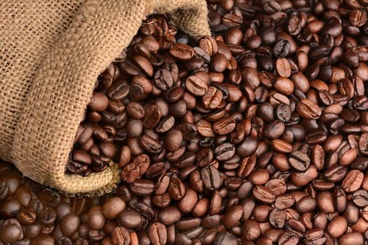 Closeup of a burlap sack of fresh roasted coffee beans spilling onto a table. The beans fill the frame. Horizontal format.