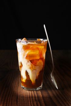 Closeup of a glass of iced coffee on a dark wood table. Fresh poured cream is permeating through the glass and a spoon is leaning on the glass. Vertical shot on a black background with copy space.