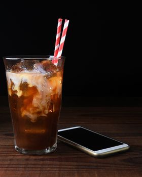 A glass of iced coffee with straws on a dark wood table. Fresh poured cream is permeating through the glass with a cell generic phone laying next to the glass. Vertical with copy space.