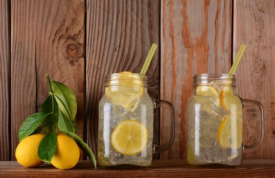 Glasses of lemonade  and lemons on a ledge in front of a rustic wooden kitchen wall. The mason jar style glasses have handles and drinking straws.
