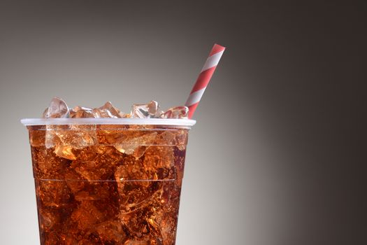 A clear plastic cup filled with ice and cola. Only half the cup is shown with a red and white striped straw over a light to dark gray background.