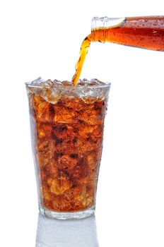 A Bottle of cola soda pouring into a glass filled with ice cubes over a white background with reflection.