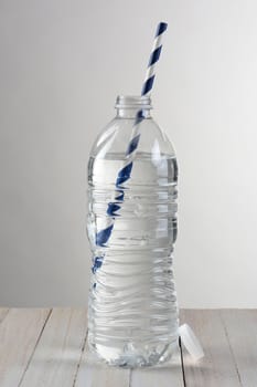 Closeup of a clear water bottle against a light to dark gray background. On a wood table the bottle has a blue and white striped drinking straw.