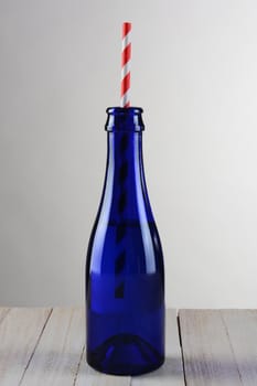 Closeup of a blue bottle with a red striped drinking straw. The bottle is on a rustic wood table with a light to dark gray background.