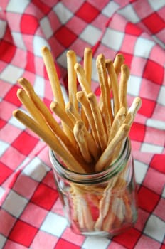 High angle closeup of bread sticks in a glass jar on a red and white checkered tablecloth. Vertical format.
