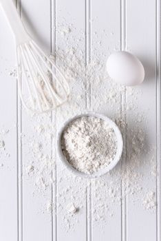 White baking still life. Top view of items for baking: eggs, flour and a whisk on a white beadboard surface. 