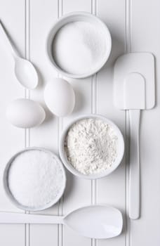 All white baking still life. Items include: spatula, whisk, eggs, flour, salt, sugar, bowls and spoons.
Vertical format shot from a high angle.